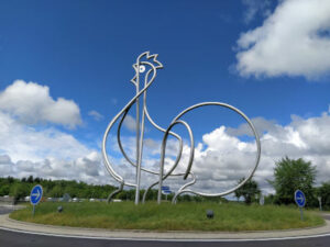 Ein Skulptur von Hahn aus Metall Röhren gemacht an einem Kreisverkehr.